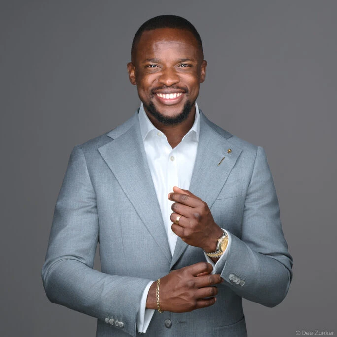 Smiling male professional in light gray suit adjusts cufflinks, studio portrait with gray background, Houston corporate headshot photography.