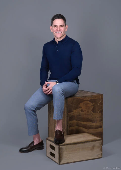 Male executive poses on wooden crate in navy polo and gray trousers, studio corporate headshot with gray background, Houston TX.