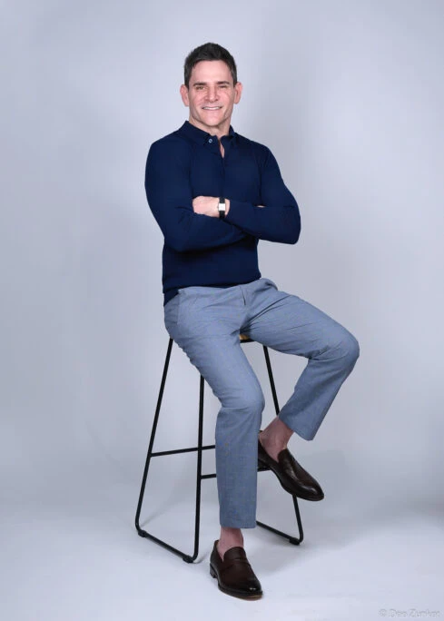 Male executive poses on stool with arms crossed in a professional Houston studio portrait against a gray background, wearing navy and gray.