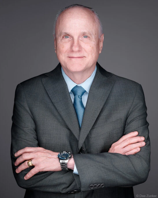 Senior male attorney in dark suit and blue tie, arms crossed, studio portrait on gray background, Houston corporate headshot photography.