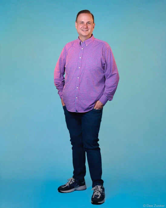 Male professional in pink checkered shirt and dark jeans poses for studio portrait against blue background in Houston.
