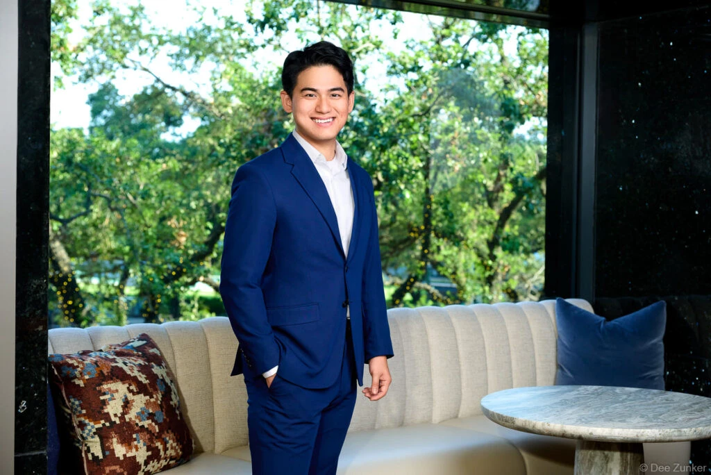 Young male professional in navy blue suit poses for environmental corporate portrait in upscale Houston interior setting with garden view.