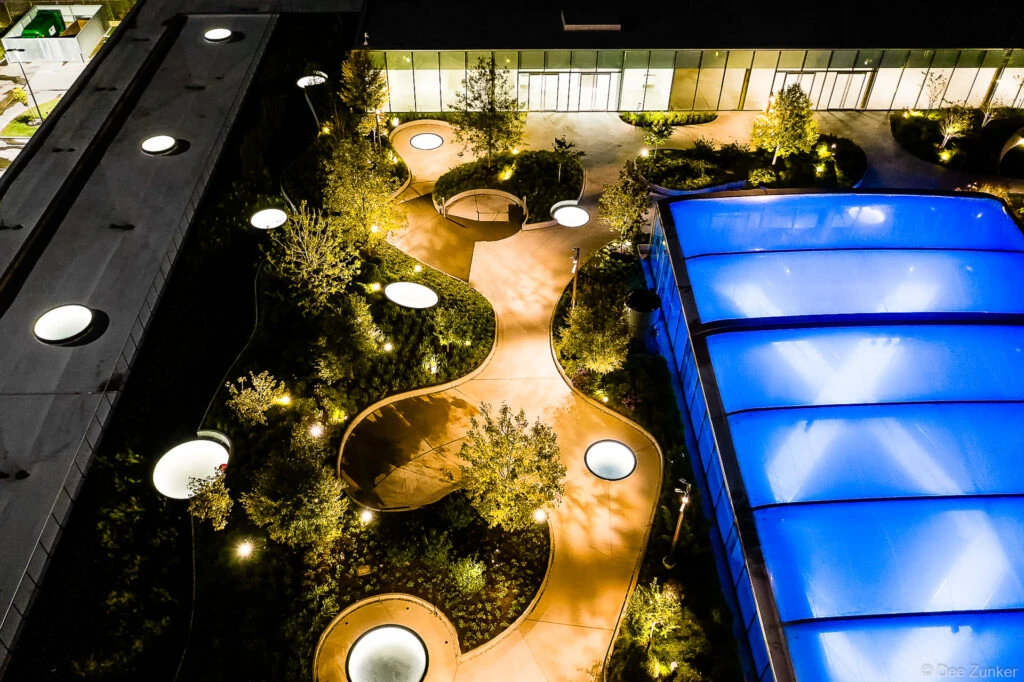 Aerial night view of Post Houston rooftop garden with illuminated curved pathways, glowing skylights, trees, and blue glass canopy.