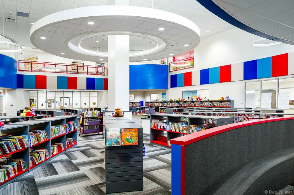 Colorful elementary school library interior featuring gray and red bookshelves, acoustic ceiling panels in blue and red, Houston TX.