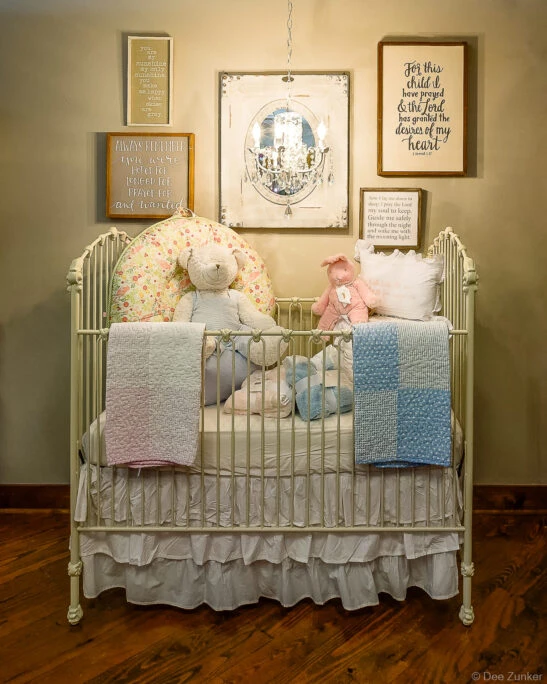 Styled baby nursery vignette featuring a white iron crib, plush toys, quilts, and inspirational wall art for retail gift product photography.