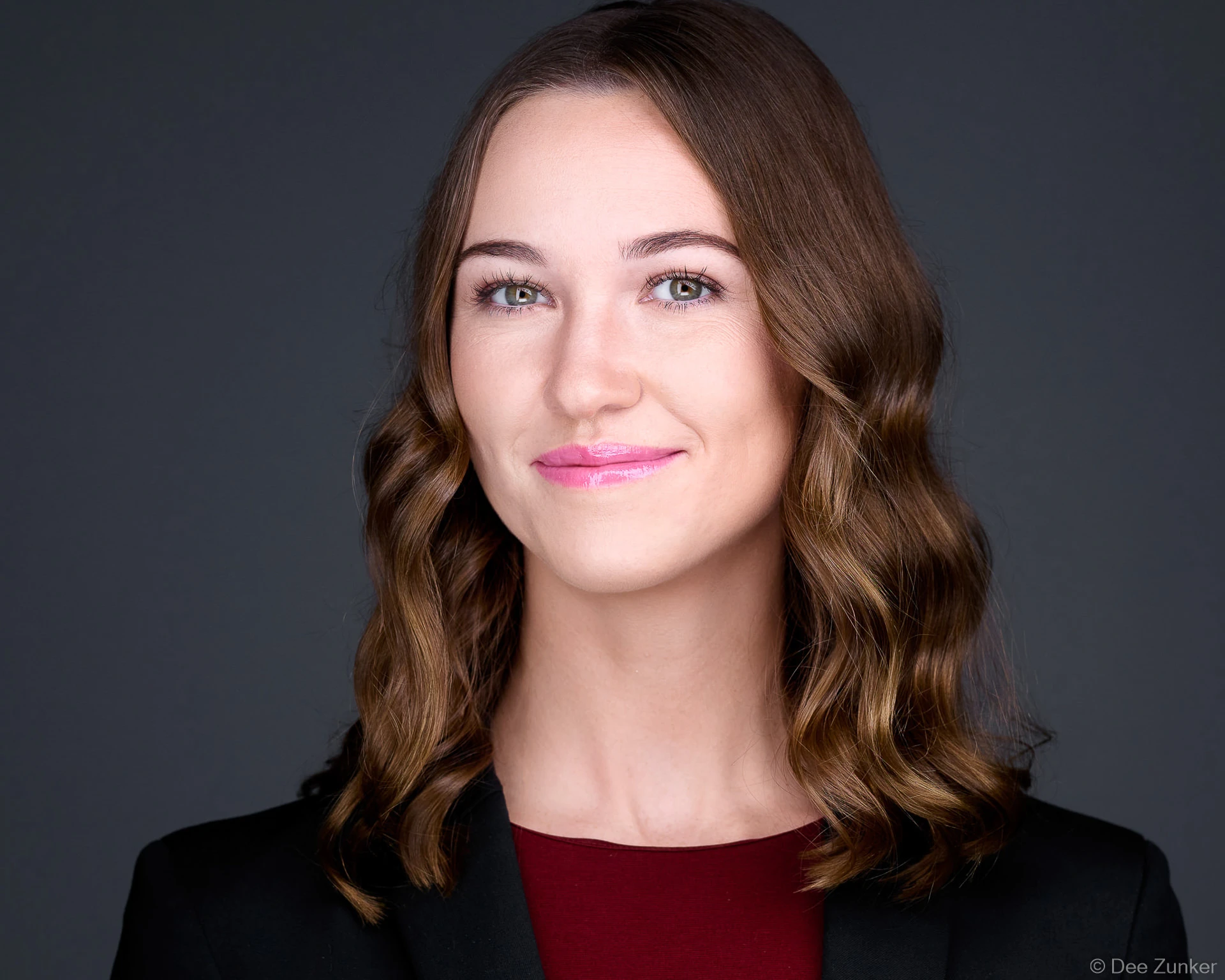 Professional woman in black blazer and burgundy top smiles confidently against a dark grey studio backdrop, Houston business headshot.