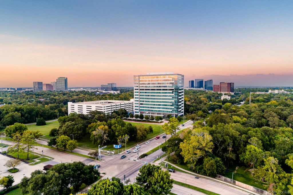 Aerial drone photo of a glass office tower at dusk along Eldridge Parkway in Houston's Energy Corridor, surrounded by tree canopy