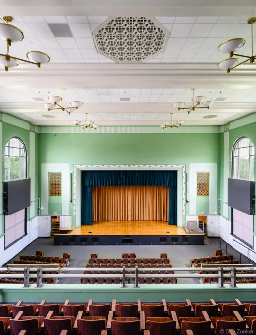 Renovated school auditorium interior with sage green walls, wood stage, teal curtains, brass chandeliers, and tiered seating in Houston