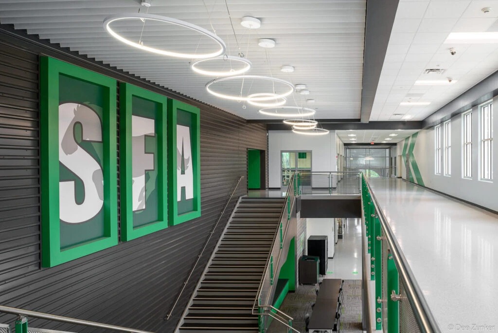 Modern school hallway with SFA lettering, circular pendant lights, staircase, and green accents at Austin High School, Houston, TX.