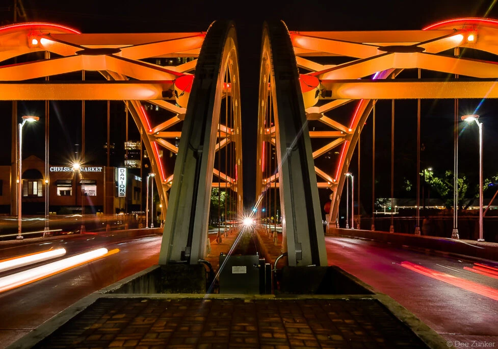 Long-exposure night photograph of the Broadway Street Bridge in Houston, TX, showcasing illuminated orange arch structure with light trails from passing vehicles.