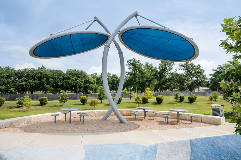 Architectural exterior of a dragonfly-shaped blue canopy shade structure with picnic tables at Clements Crossing park, Houston TX.
