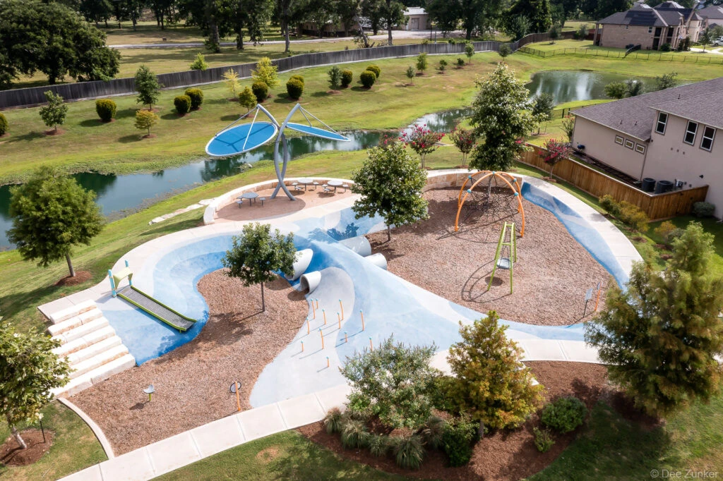 Aerial drone photo of a modern playground at Clements Crossing featuring blue sculptural surfaces, climbing structures, and a pond, Houston TX.