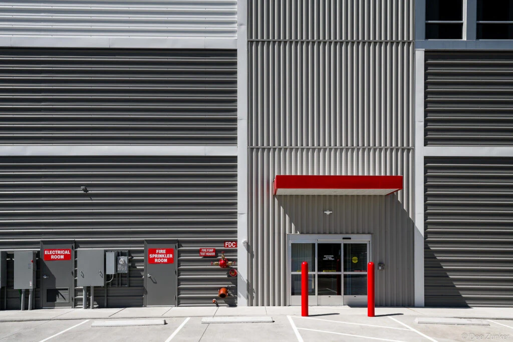 Exterior facade of a modern storage facility featuring corrugated metal cladding, red accents, electrical room and fire sprinkler signage, and glass entry doors