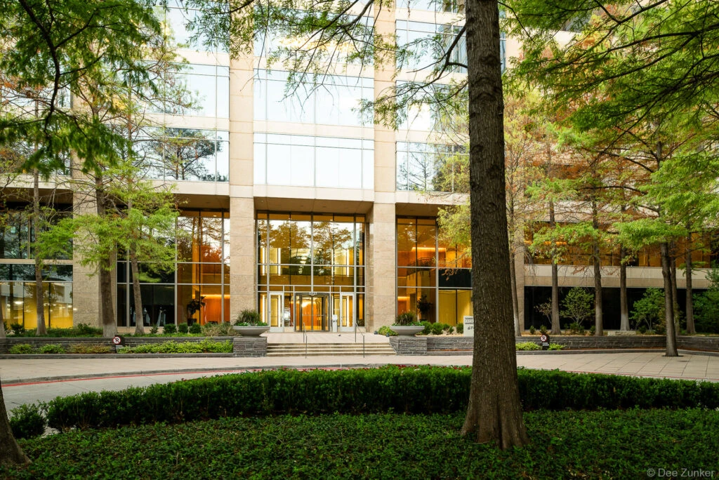 Modern glass and limestone office tower entrance with manicured landscaping and tall trees at a Dallas corporate campus, exterior architectural photography.