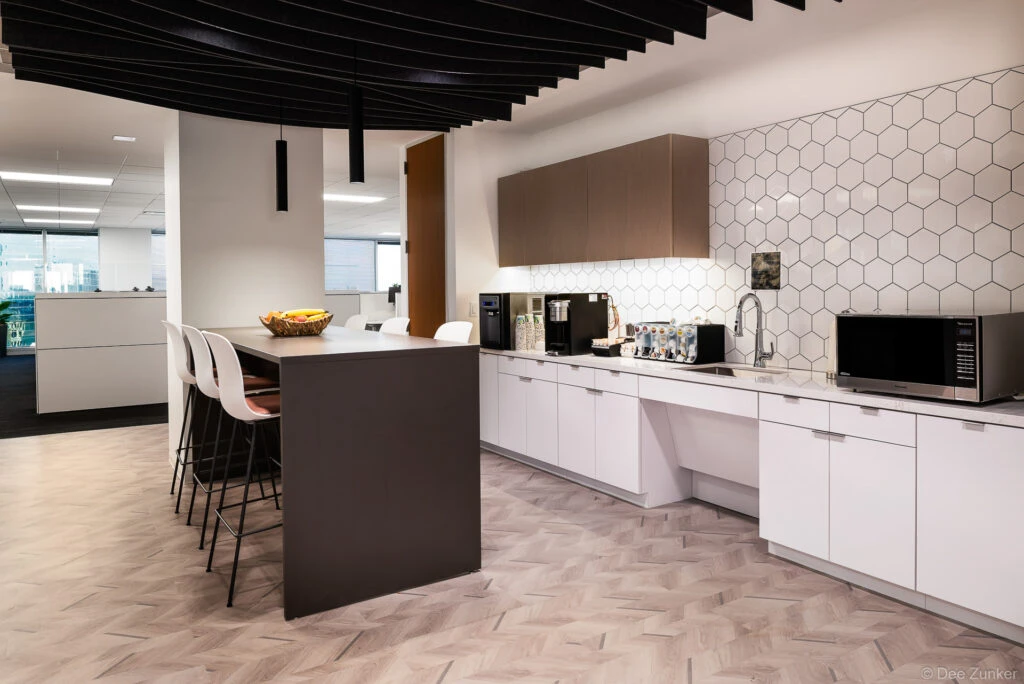 Modern office break room with white hexagonal tile backsplash, herringbone wood floors, dark island with bar seating, and coffee station in Dallas.