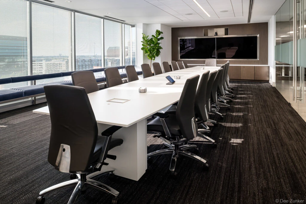 Modern executive conference room with long white table, leather chairs, large display screen, and floor-to-ceiling windows overlooking Dallas skyline