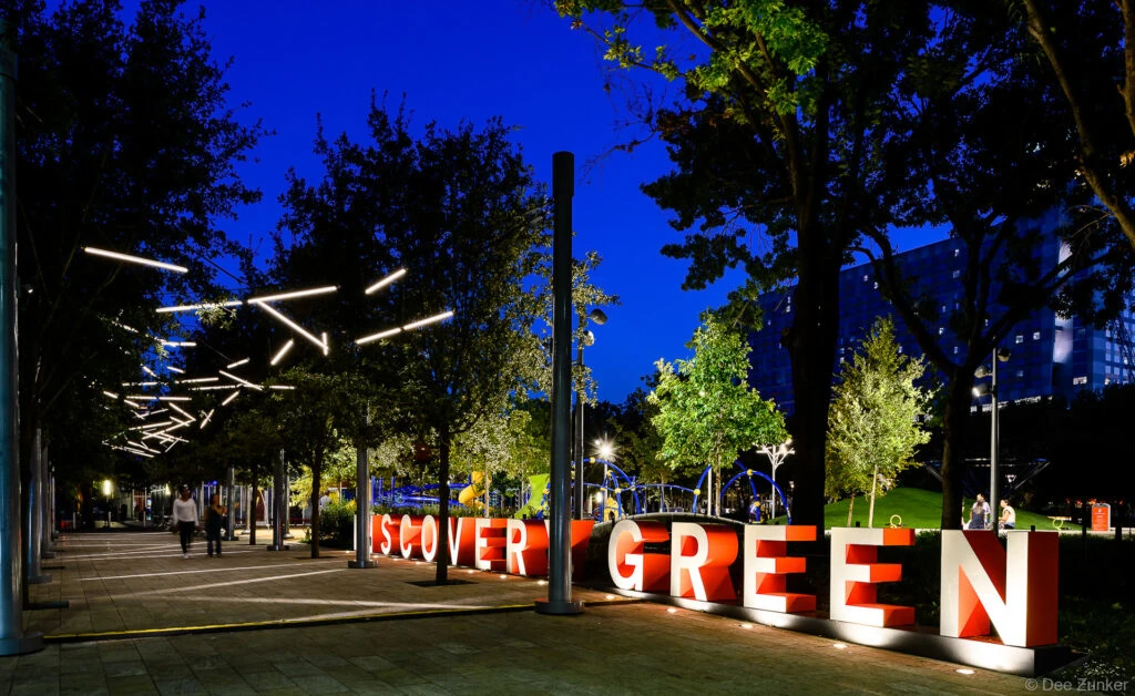 Large red Discovery Green sign illuminated at night with geometric LED light installations along a tree-lined promenade in Houston, TX.