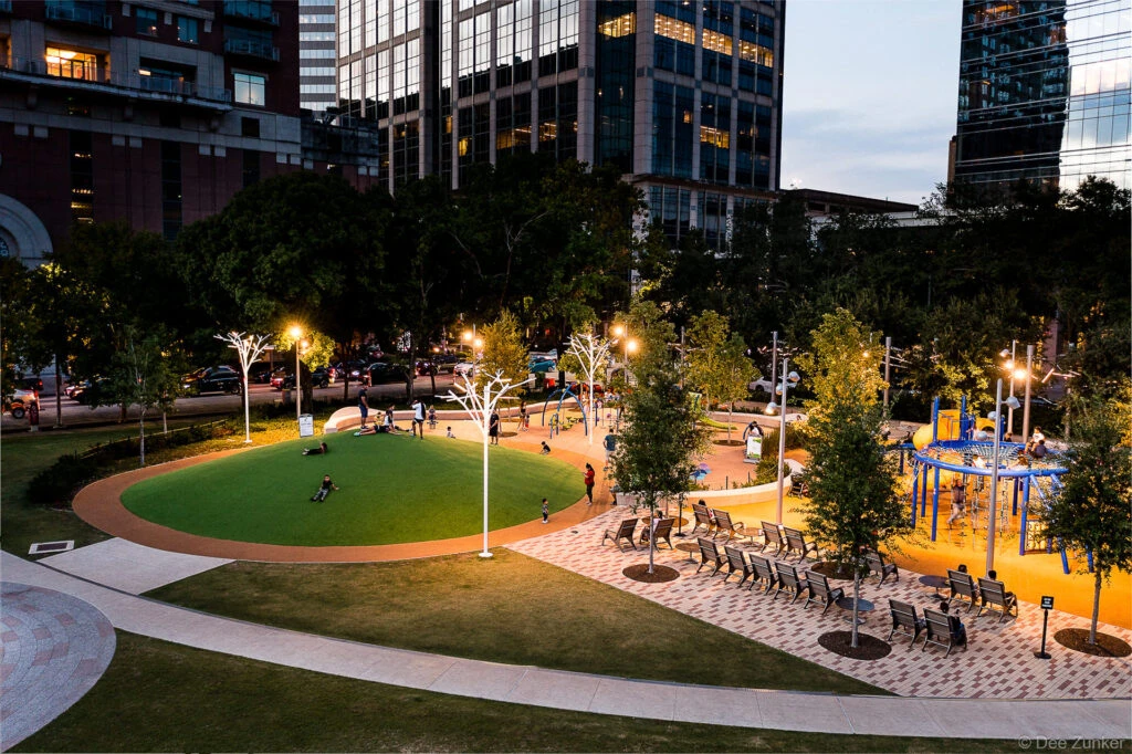 Aerial dusk view of Discovery Green park in Houston showing illuminated sculptural light trees, playground, and green lawn surrounded by downtown skyscrapers.
