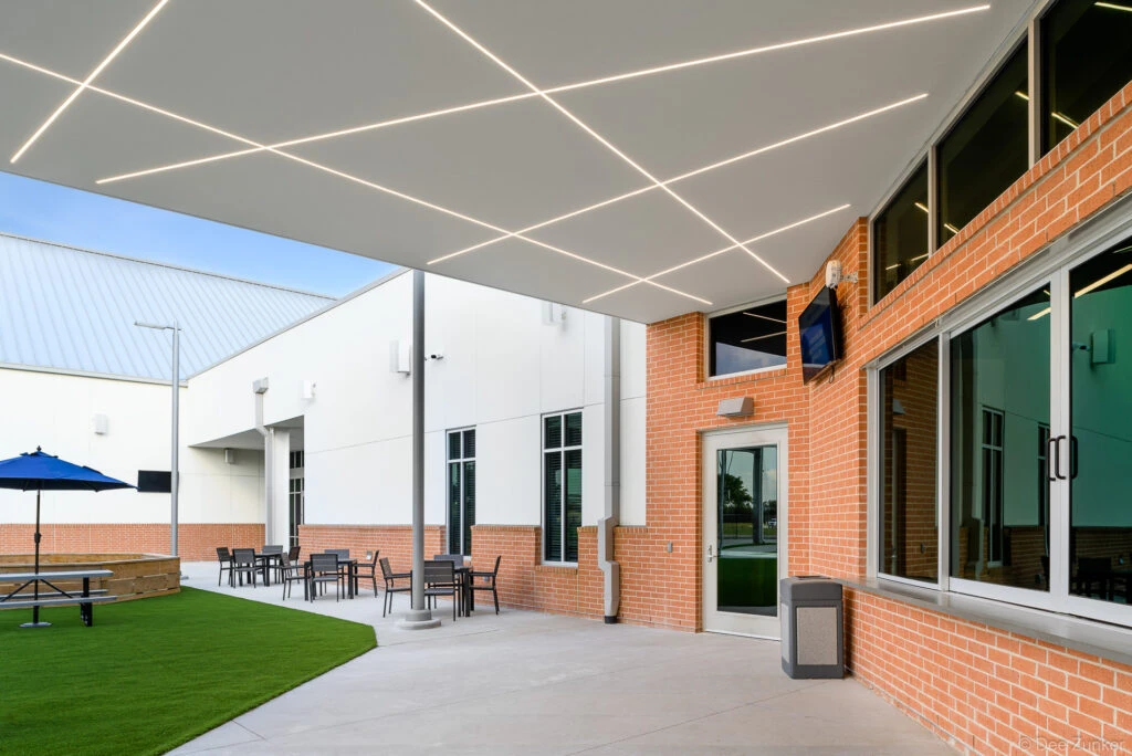 Covered outdoor courtyard of a church student center featuring geometric LED ceiling lighting, brick exterior, patio seating, and artificial turf in Spring TX