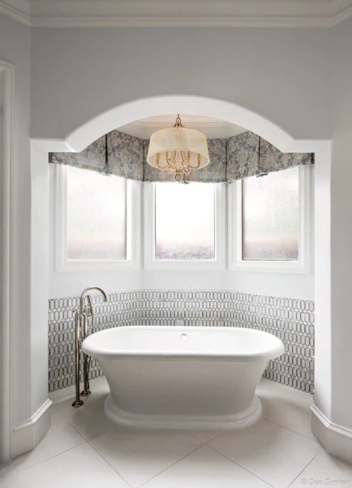 Elegant white freestanding soaking tub in an arched alcove with mosaic tile surround, chandelier, and bay windows in a Houston interior.