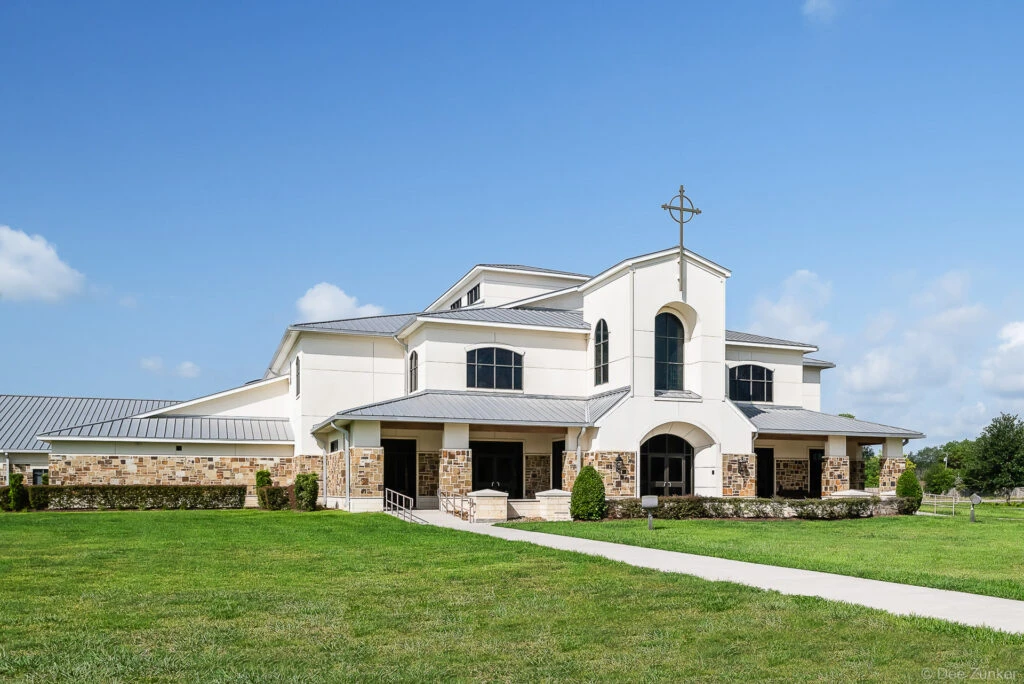 Exterior of a modern white stone church with a cross tower, metal roof, and manicured lawn on a clear sunny day in Friendswood Texas
