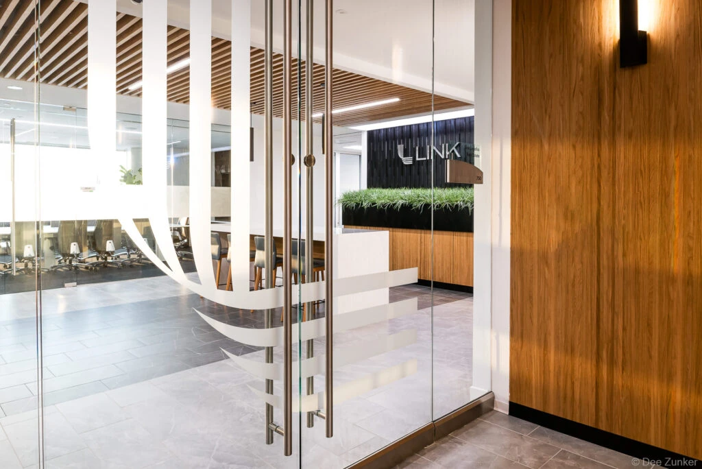 Glass door entrance to a modern Dallas corporate office lobby featuring wood paneling, reception desk with greenery, and slatted ceiling details.