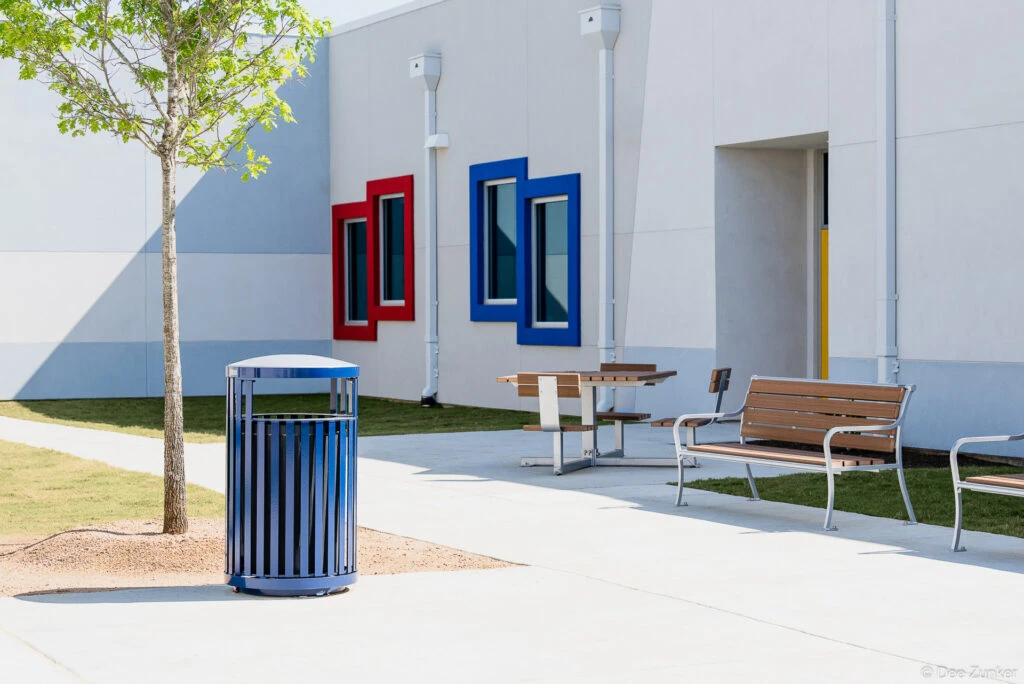 Exterior courtyard of Harmony Spring school featuring blue trash bin, wood-slat benches, picnic table, young tree, and colorful window frames.