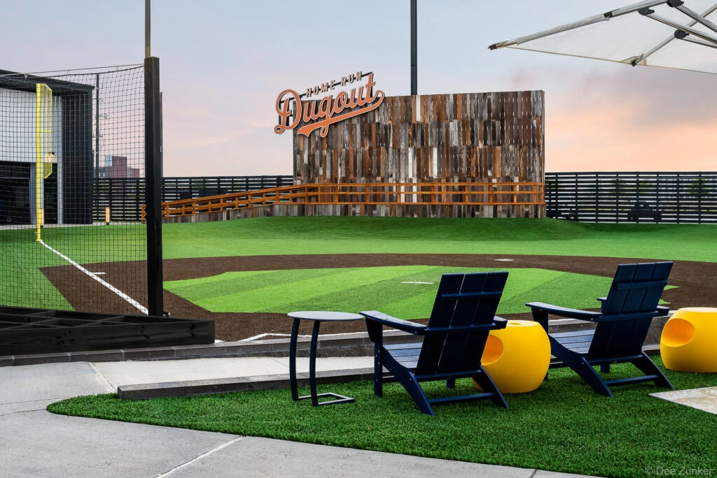 Exterior architectural view of Home Run Dugout Katy featuring artificial turf baseball field, Adirondack chairs, and reclaimed wood backdrop at dusk.