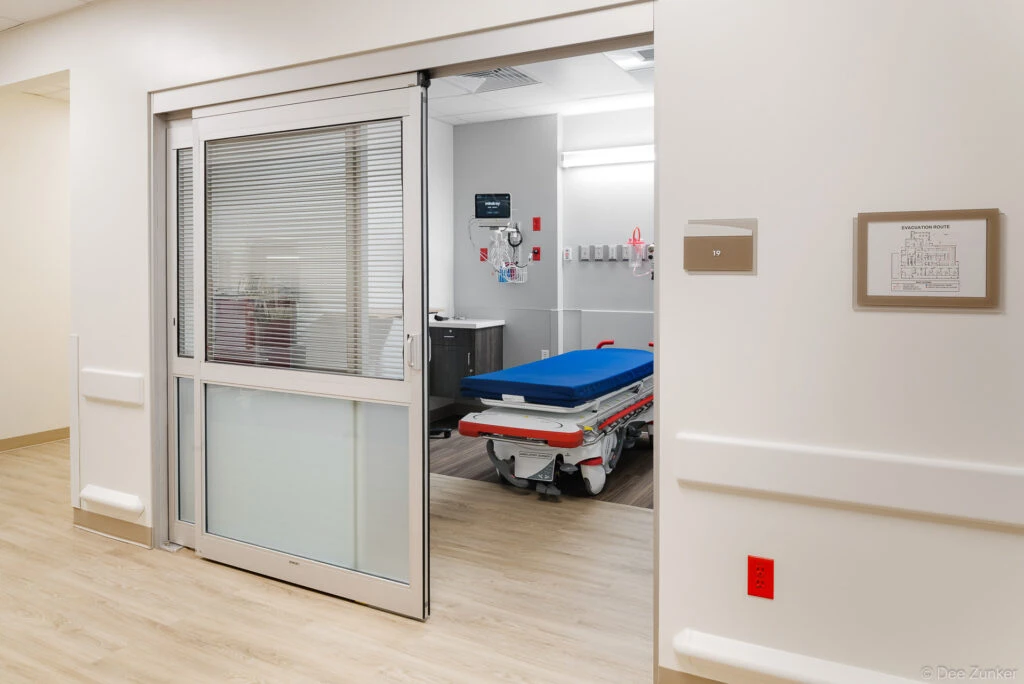 Modern hospital emergency room bay interior showing a blue medical gurney, patient monitoring equipment, and sliding glass privacy door in a Houston healthcare facility.