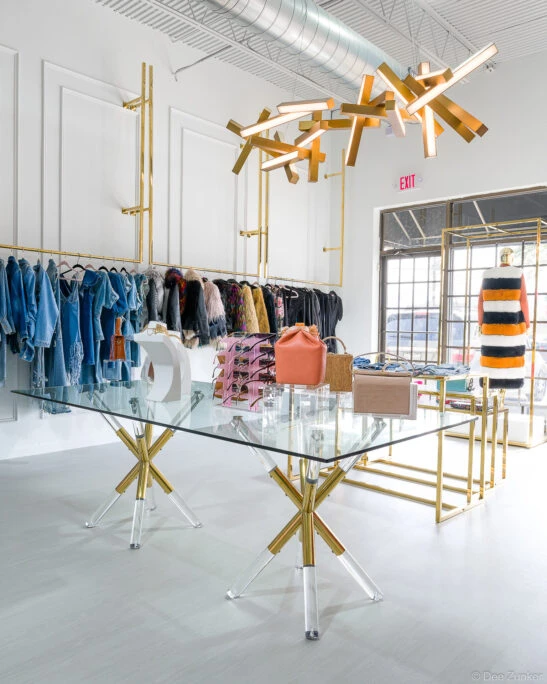 Upscale Houston boutique interior with glass display table, gold lighting fixture, handbags, sunglasses, and clothing racks with denim and fur coats.