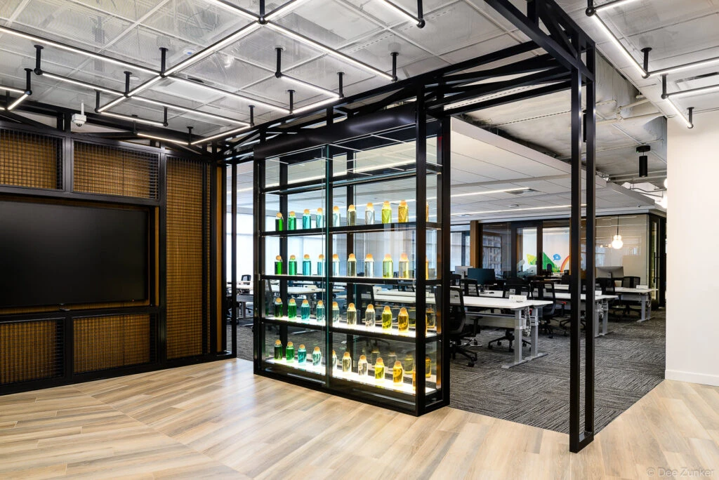 Modern corporate office interior featuring a glass display shelf with colorful bottles, open workstations, wood accents, and industrial ceiling lighting in Houston