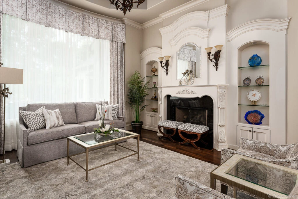 Formal Houston living room with white ornate fireplace, gray sofa, leopard print ottomans, built-in shelving, and tall windows with patterned drapes.