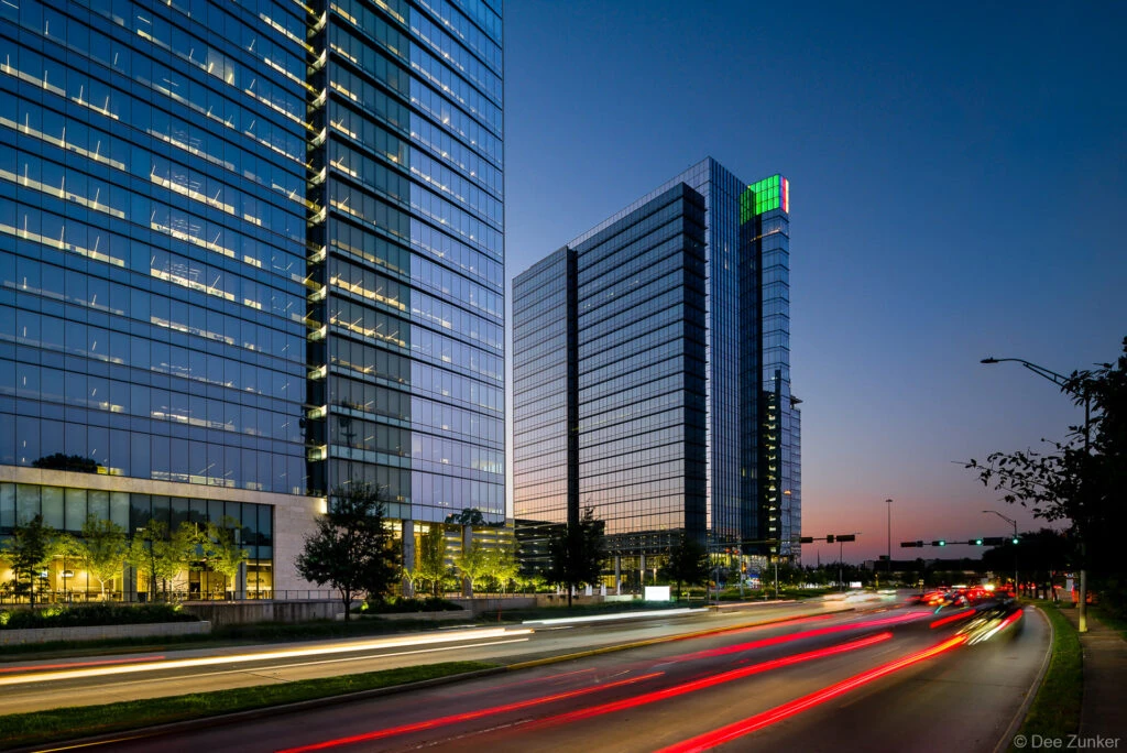 Glass office towers in Houston Energy Corridor at twilight with colorful light trails streaking along a busy boulevard