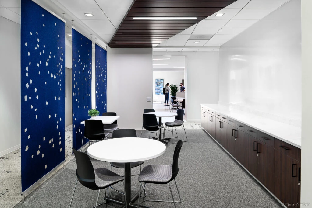 Modern office breakroom with round white tables, black chairs, blue perforated acoustic panels, and dark wood cabinetry in Houston institutional renovation