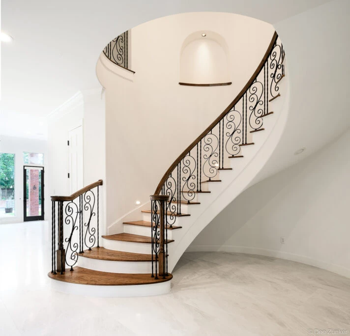 Elegant curved staircase with wrought iron scroll balusters, wood handrail, and white risers in a luxury Houston residential interior.