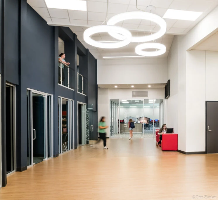 Two-level college student center interior hall with circular pendant lights, wood floors, red seating, and a glass-walled learning hub in Baytown TX