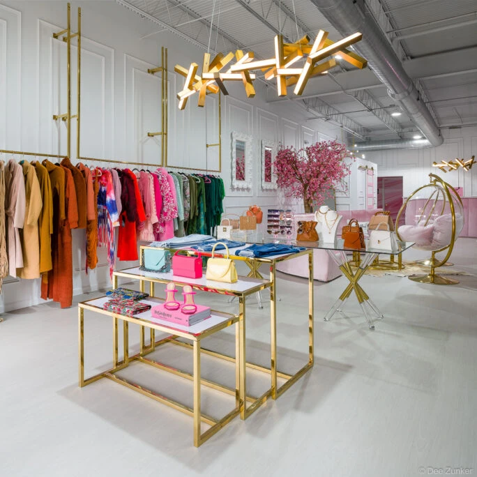 Upscale Houston boutique interior with gold display tables, colorful clothing racks, designer handbags, and pink decor accents in a modern retail space.