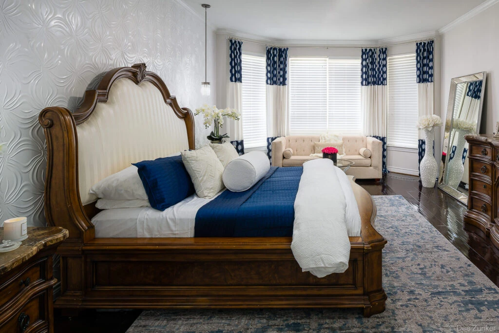 Ornate king bed with navy and white bedding in a luxury Houston master bedroom featuring bay windows and tufted seating area.