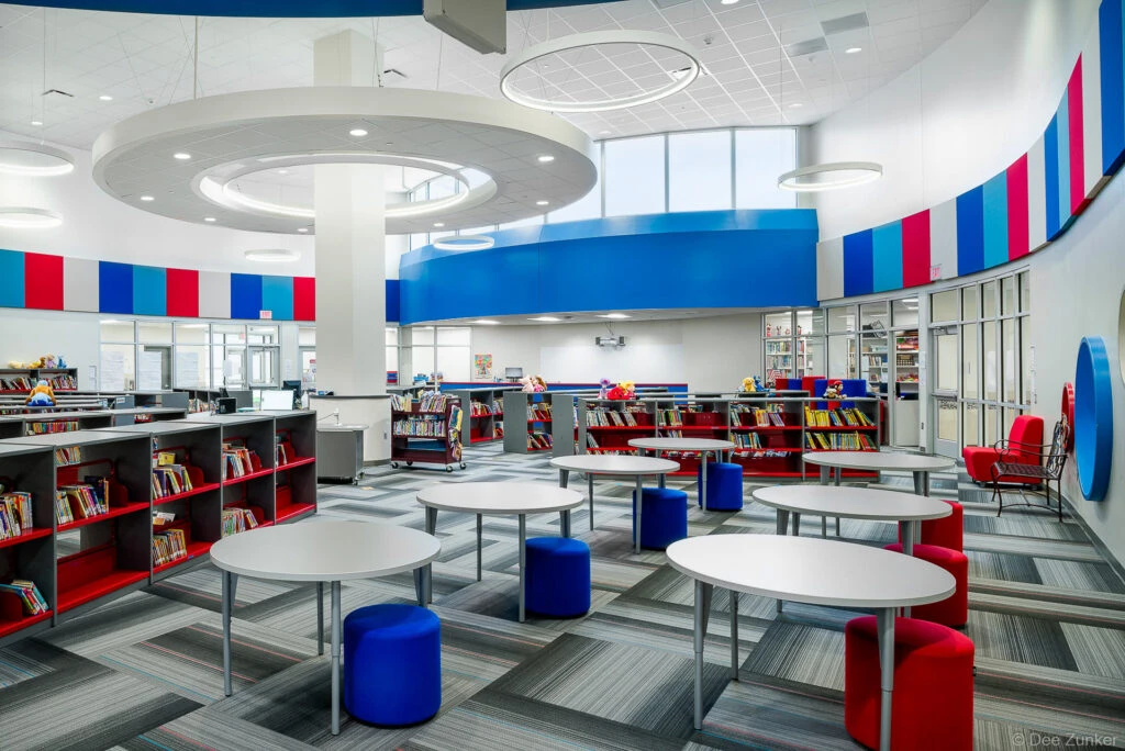 Bright elementary school library interior with round tables, colorful red and blue seating, bookshelves, and modern circular ceiling lights in Houston