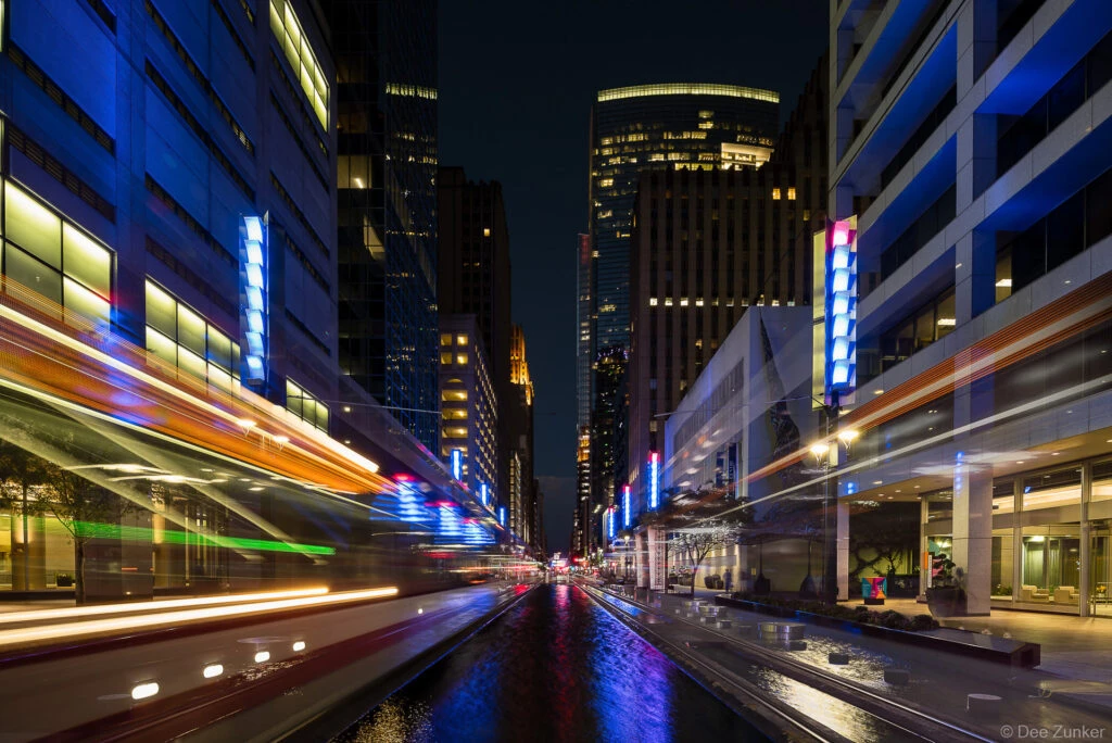 Long exposure night photograph of Main Street Square Houston with light trails from passing trains and glowing blue facade lighting