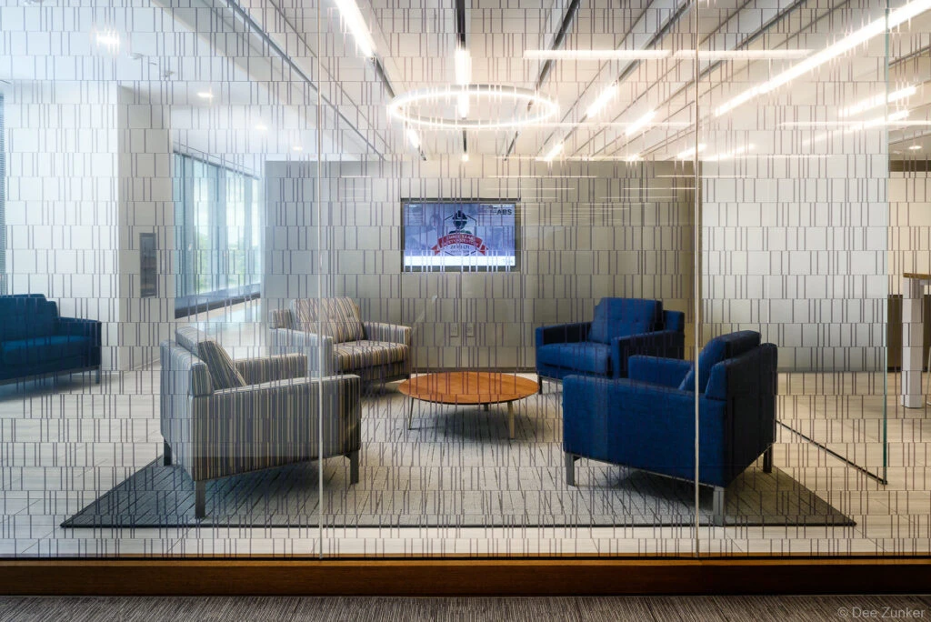 Modern corporate lounge viewed through patterned glass partition, featuring blue and gray armchairs, wood coffee table, and wall-mounted display screen