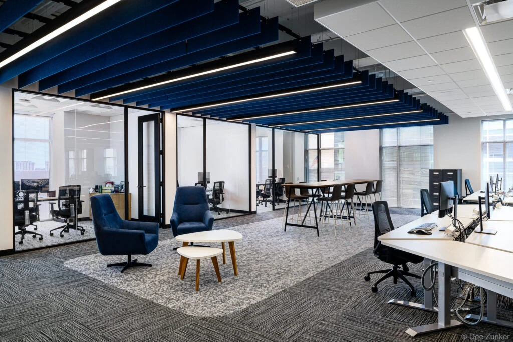 Modern open-plan corporate office featuring blue lounge chairs, collaborative workstations, glass partition walls, and bold blue acoustic ceiling baffles, Houston TX