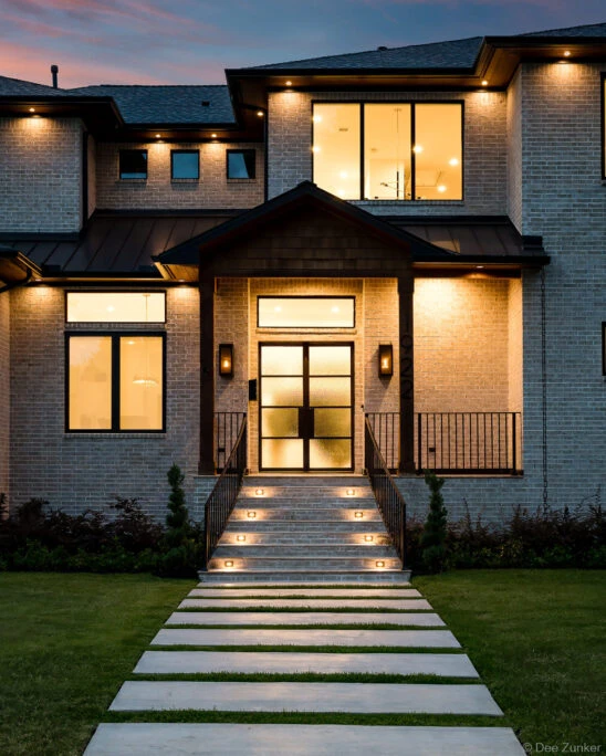 Illuminated modern brick home exterior at dusk with lit staircase, black-framed glass entry door, and manicured lawn in Houston TX.