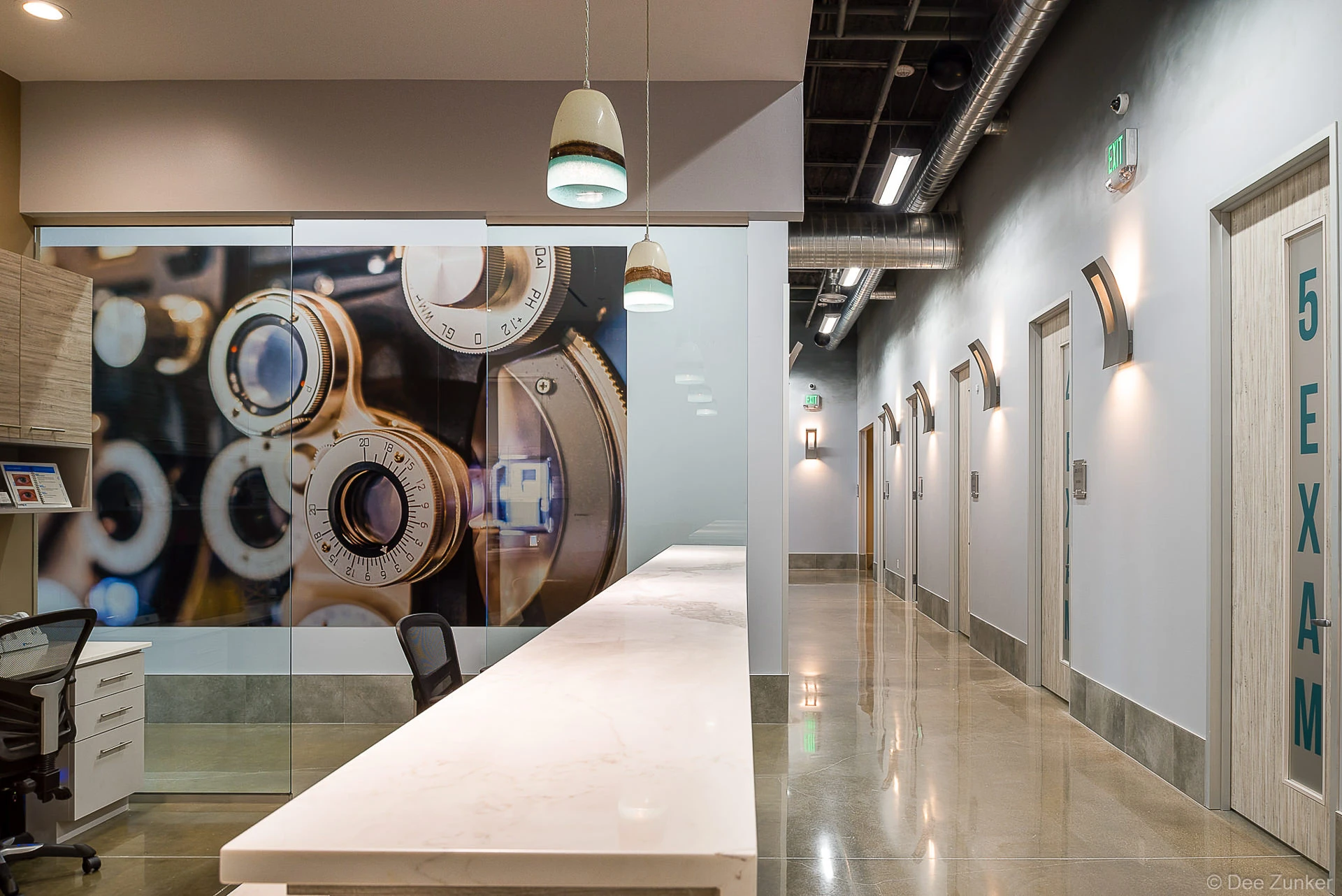 Modern optometry clinic hallway with exam rooms, marble reception counter, phoropter wall mural, and industrial-style lighting in Houston.