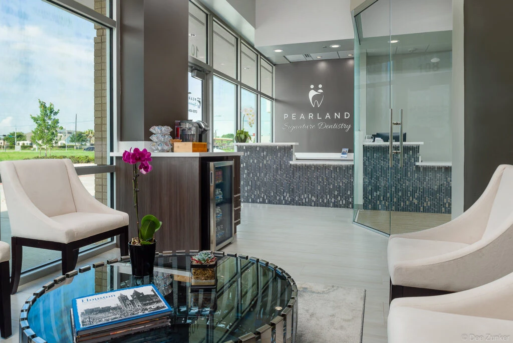 Modern dental office reception area with mosaic tile front desk, white lounge chairs, orchid centerpiece, and glass walls in Pearland TX