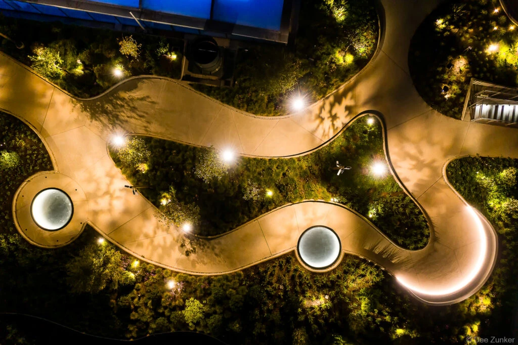 Aerial night drone view of POST Houston rooftop garden with winding illuminated walkway, glowing skylights, and lush plantings