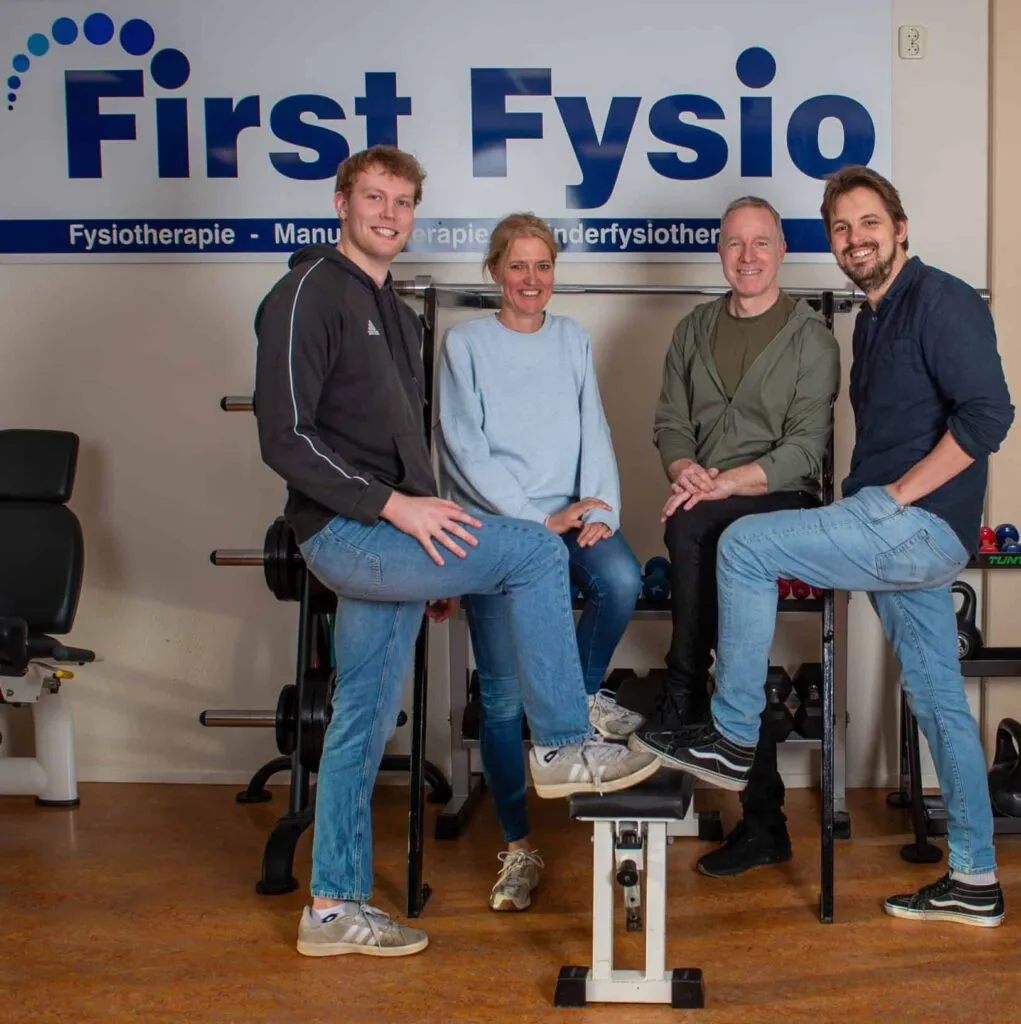 Vier mensen glimlachen en poseren samen in een fysiotherapiekliniek, voor een bord met de tekst First Fysio. Op de achtergrond zijn fitnessapparaten te zien.