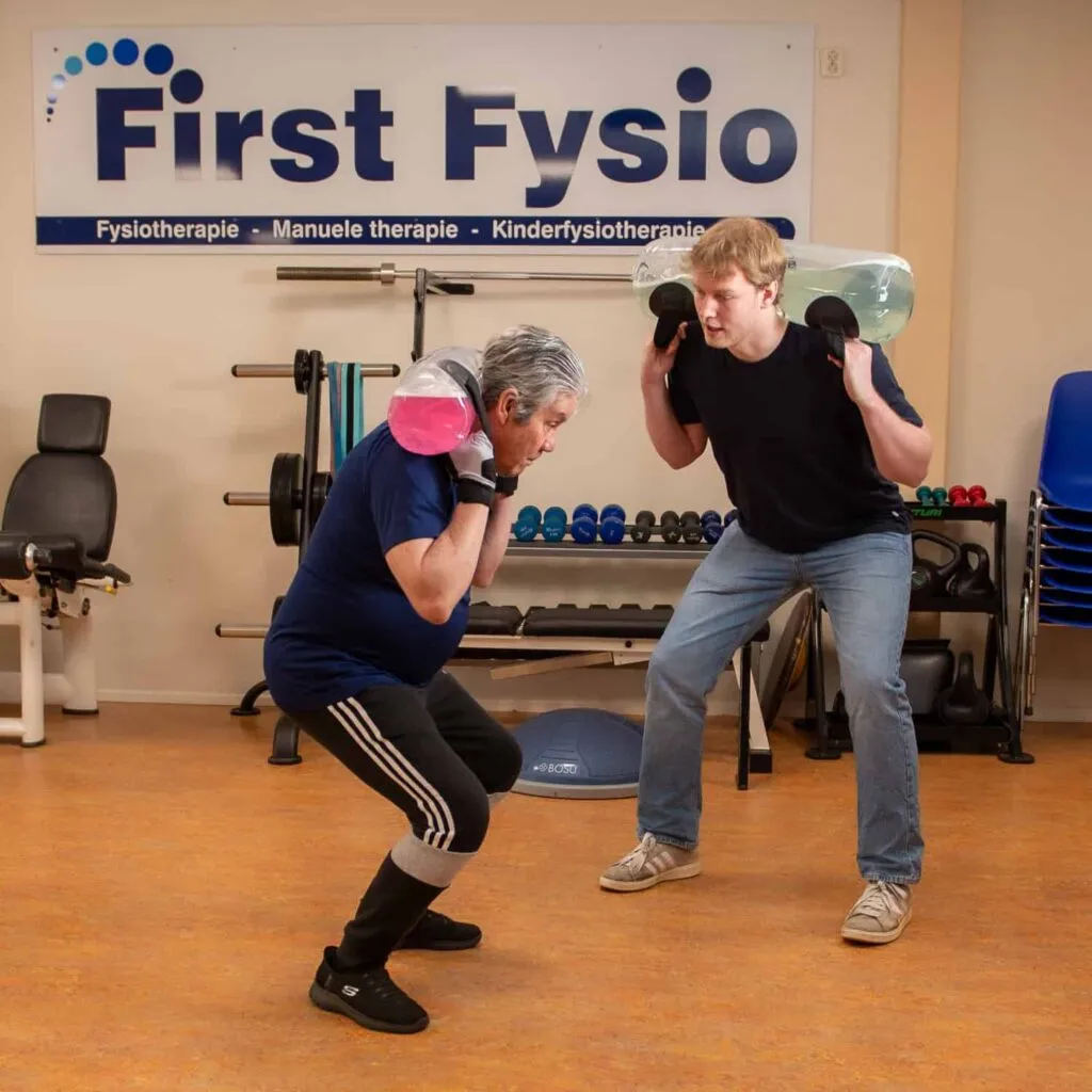 Twee mensen oefenen in een sportschool, ze hurken terwijl ze gewichten op hun schouders houden. Op een bord achter hen staat First Fysio. Fitnessapparatuur en gestapelde stoelen zijn zichtbaar op de achtergrond.
