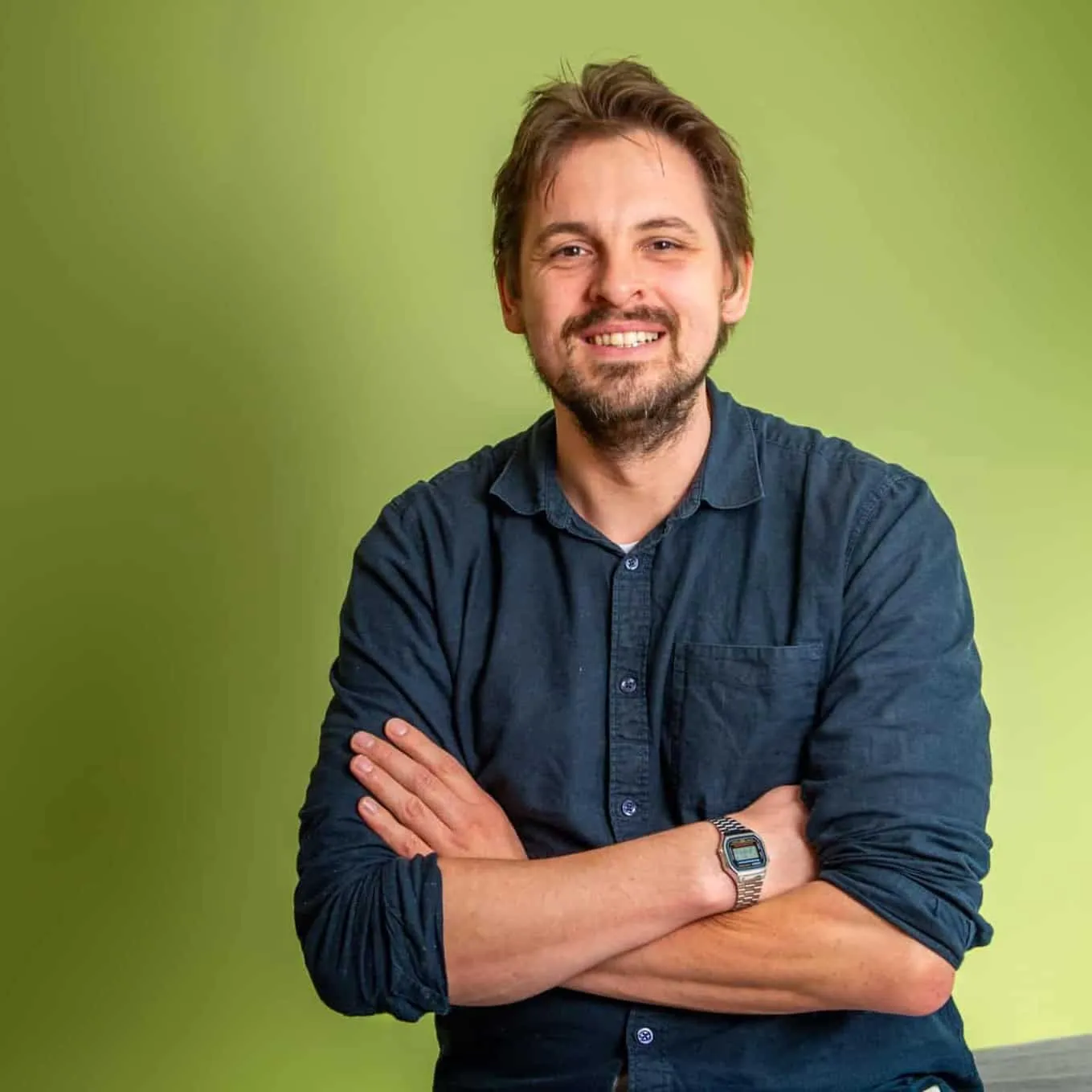 Een man met bruin haar en een baard, een donkerblauw overhemd en een digitaal horloge, glimlacht met zijn armen over elkaar voor een lichtgroene achtergrond.