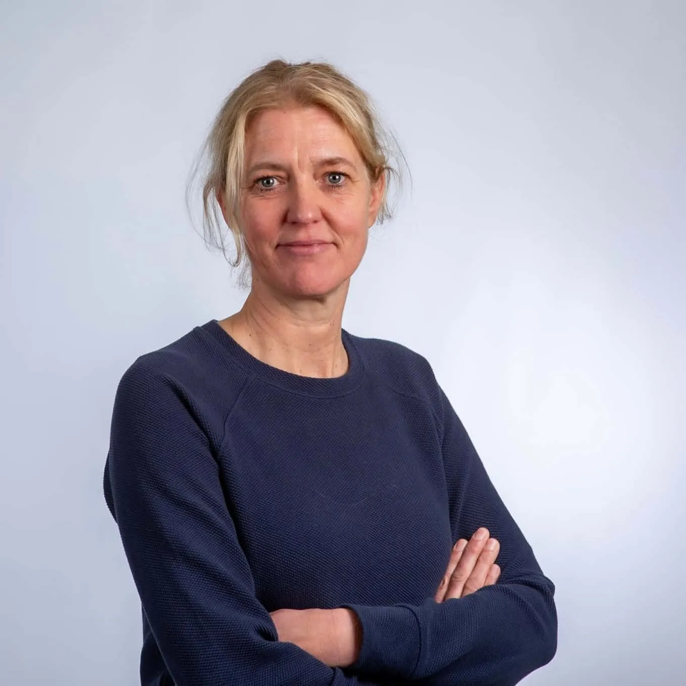 Een vrouw met achterover gebonden blond haar staat met haar armen over elkaar, draagt een donkerblauwe trui en glimlacht lichtjes tegen een effen lichte achtergrond.